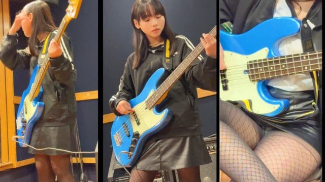黒いジャージと黒いスカート、網タイツ姿の人物が、黄色い肩ひもで青いベースを掛け、立ったまま片手でネックを押さえ、もう片方で弦を弾いている。