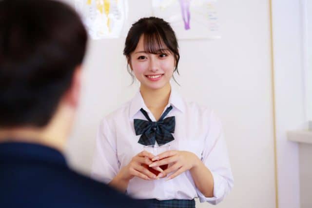 白いシャツとチェック柄のリボン姿の女性が、透明なカップを両手で持って微笑み、向かいに座る人物へ顔を向けている。髪は後ろでまとめられている。